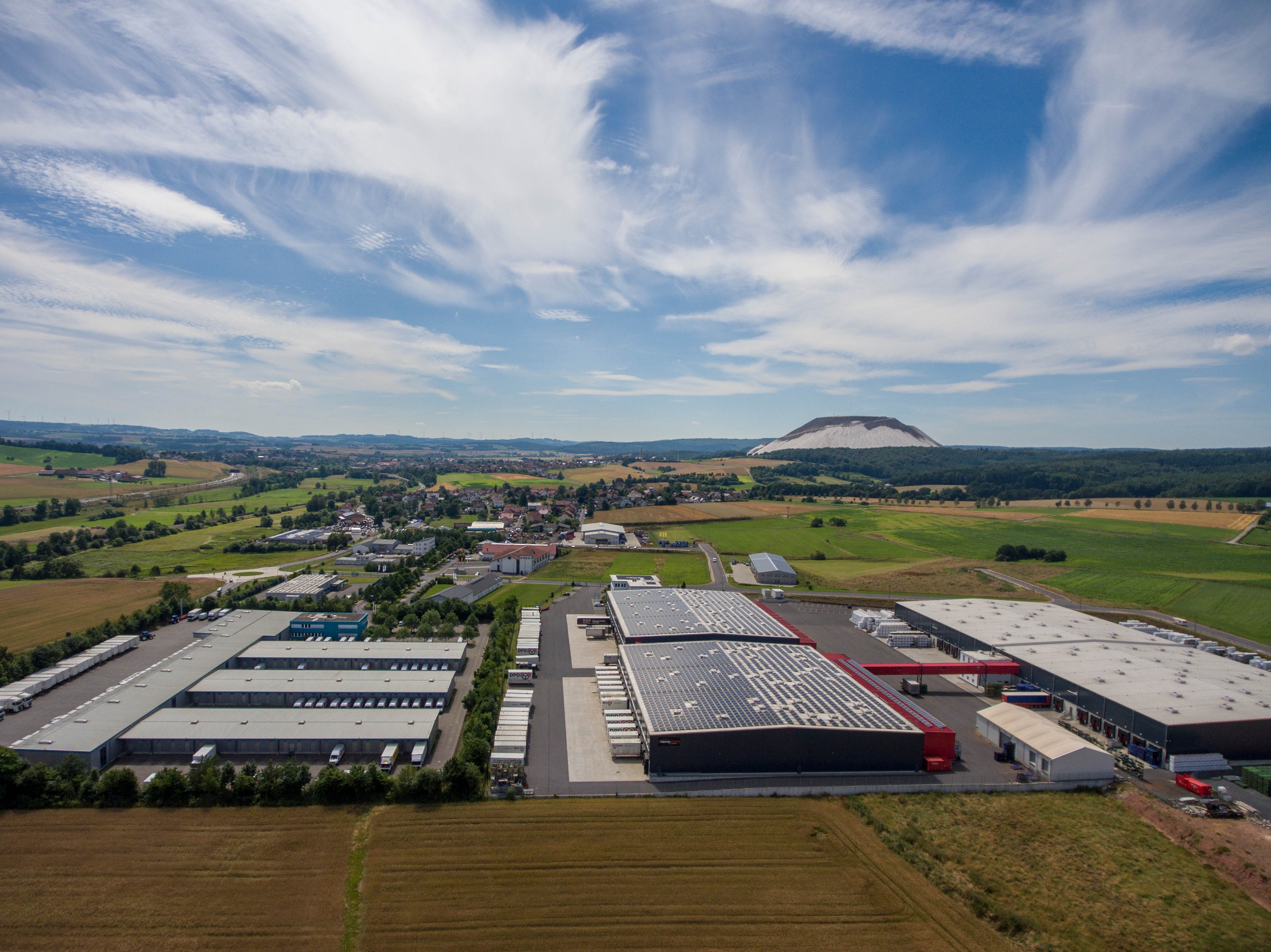Luftaufnahme bei Tageslicht - im Vordergrund sind Unternehmensgebäude sowie Häuser zu sehen. Im Hintergrund ist ein Kaliberg zu sehen.