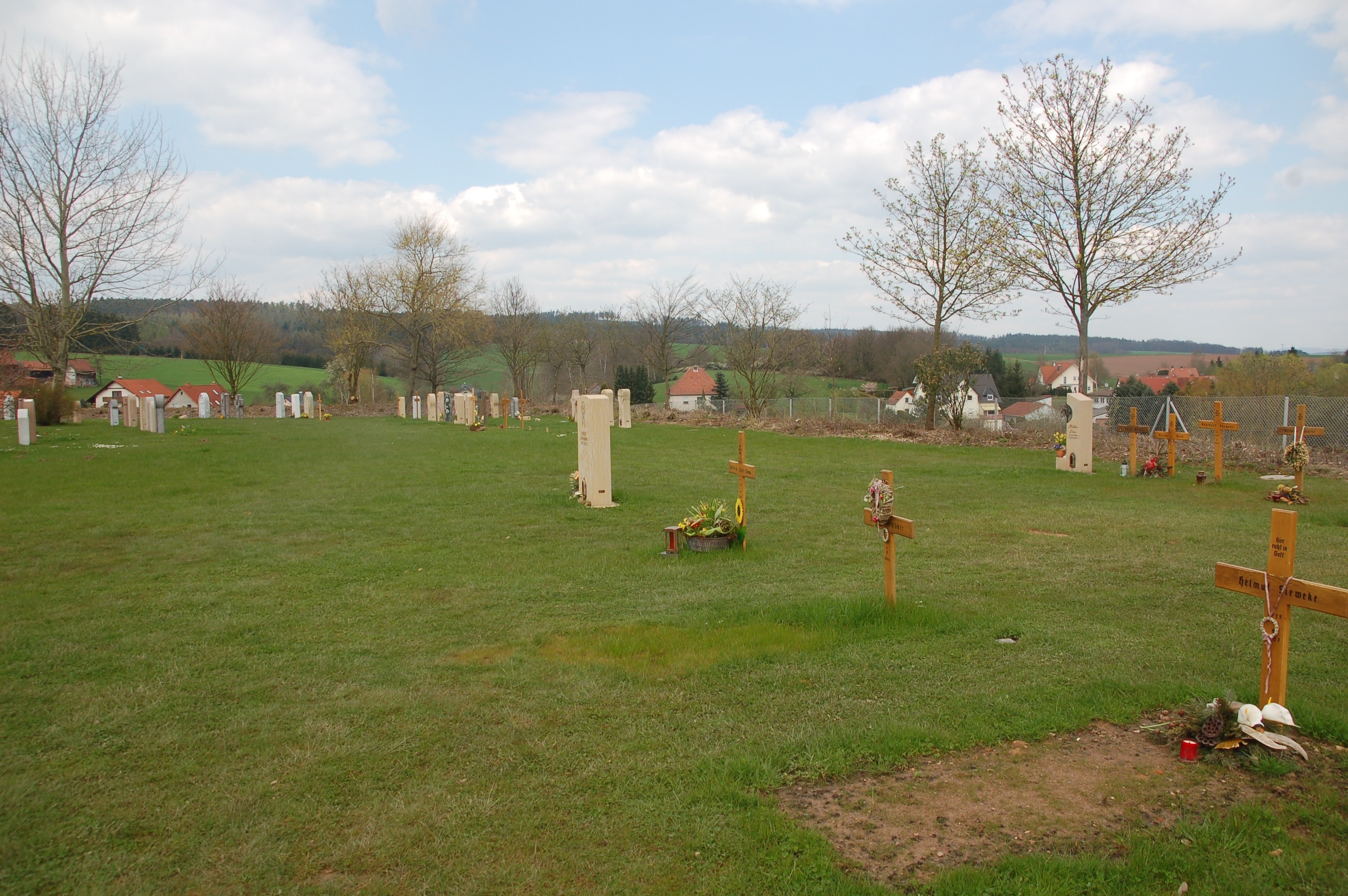 Zu sehen sind Rasengräber auf einem gemeindlichen Friedhof. Auf den Grabstätten befinden sich teilweise Grabsteine oder Grabkreuze. Rundherum befindet sich Wiese.