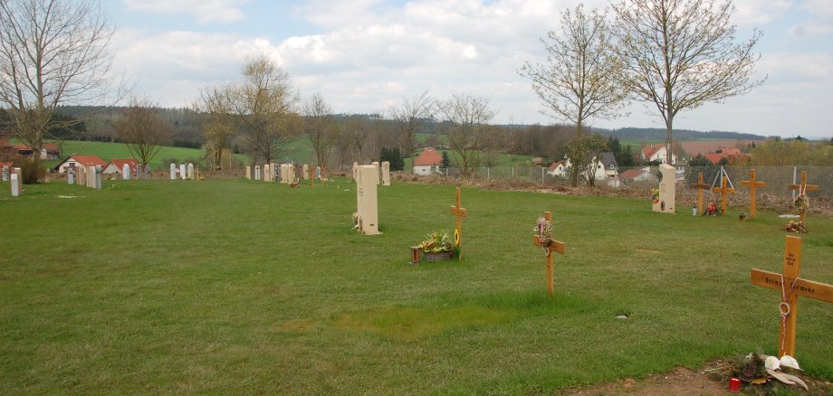 Zu sehen sind Rasengräber auf einem gemeindlichen Friedhof. Auf den Grabstätten befinden sich teilweise Grabsteine oder Grabkreuze. Rundherum befindet sich Wiese.
